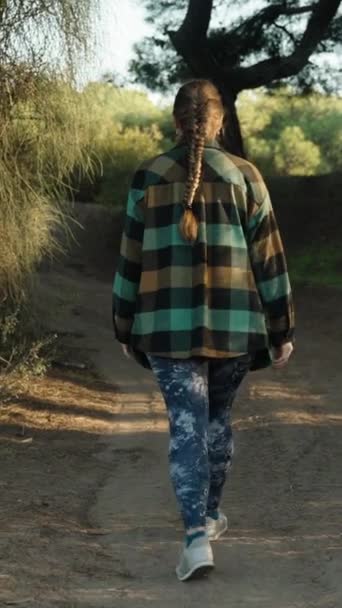 平らなシャツと青いレギンスの若い女性は日没時に森を歩き 木の葉に触れ 後ろの景色を眺めます 垂直ビデオ ソーシャルメディア — ストック動画
