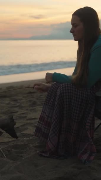 海で日没する若い女性は 火事でマシュマロを揚げる 寒いし毛布で覆われている 垂直ビデオ ソーシャルメディア — ストック動画