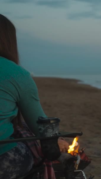 若い女性は海岸のキャンプファイヤーで毛布と揚げマシュマロで覆われた椅子に座っています 垂直ビデオ ソーシャルメディア — ストック動画