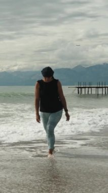 Tombul genç bir kadın, deniz kıyısında, dağların ve gök gürültülü bulutların arkasında ağır çekimde yürüyor. Dikey video Sosyal Ortamı