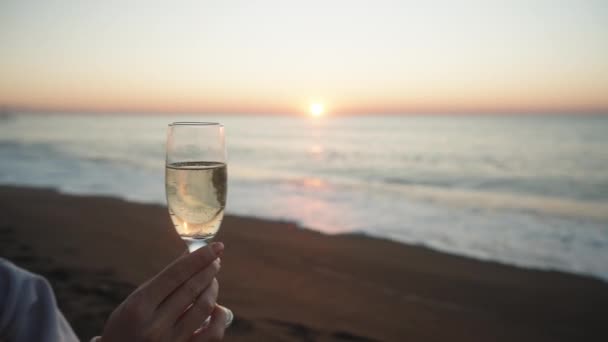 クールな朝 フーディーの若い女性が海と昇る太陽の背景に対してシャンパンのグラスを保持 — ストック動画