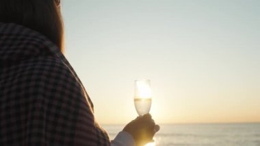 Battaniyeye sarılı genç bir kadın elinde bir bardak şampanyayla Denizde Gündoğumuyla karşılaşıyor..