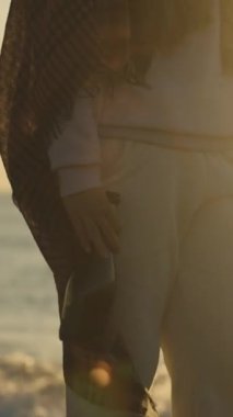 Sıcak giyimli genç bir kadın elinde bir şişe şampanyayla deniz kıyısında duruyor. Güneş ve suyun üzerindeki parıltı geride bırakıldı. Dikey video Sosyal Ortamı