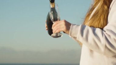 Genç bir kadın deniz kıyısındaki bir bardağa şampanya dolduruyor. Gün doğumu. Yavaş çekim.