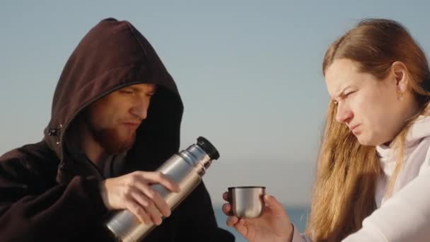 海で涼しい朝 黒いフードの男は熱湯から若い女性の温かいお茶をプール — ストック動画