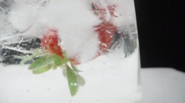 Soda bardağındaki çilek ve buz küpleri döner ve kabarcıklar yükselir, siyah zemin üzerinde makro.