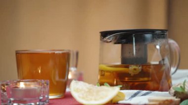 Masada mumlar yanıyor ve içinde papatya çayı ve nane çayı olan bir çaydanlık var. Limon ve zencefil yan yana yatıyorlar. Kırmızı masa örtüsü.
