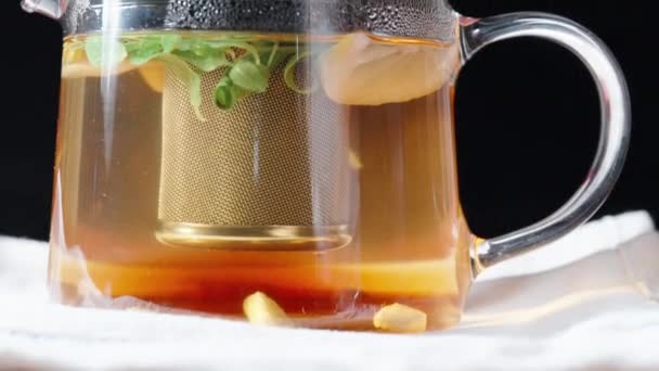 茶壶中装有金属筛子的玻璃茶壶 在白色桌布和黑色背景上 带有薄荷糖和生姜的柠檬是漂浮在体内的 多利滑行特写 — 图库视频影像