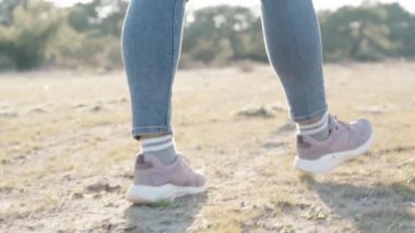 Spor ayakkabılı bir kadın kuru çimlerde ve kumda yürür. Güneşin parlak ışınlarının arka planında, aşağıdan yavaş çekimde yakın çekim.