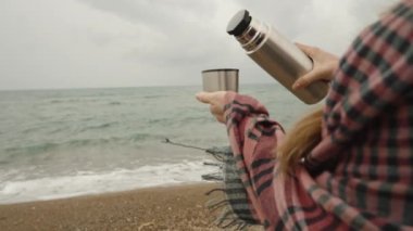 Sahilde serin bir günde ekose giymiş bir kadın termostan sıcak çay döküyor, ağır çekimde..
