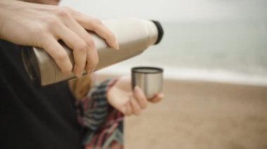 Ekoseli genç bir kadın, deniz sahilindeki termostan çay dolduruyor, yakın çekim, yavaş çekim..