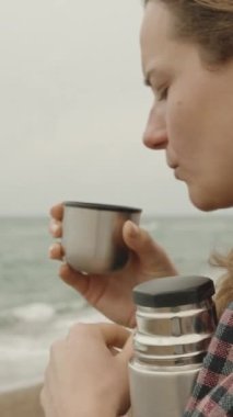 Dikey video. Deniz kenarında serin bulutlu bir gün. Ekoseli bir kız termostan sıcak çay içiyor..
