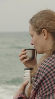 Dikey video. Genç bir kadın serin bir günde denize bakar ve termostan çay içer..