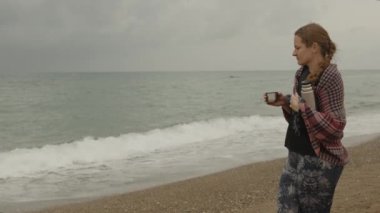 Denizde bulutlu bir gün, genç bir kadın termostan bir kupa çay tutuyor. Dalgalara bak..