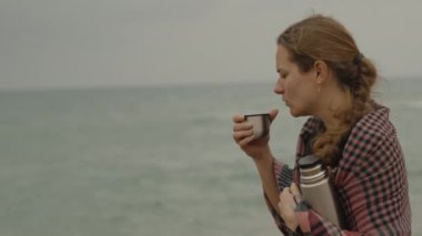 Denizde bulutlu ve rüzgarlı bir günde, genç bir kız termostan gelen sıcak çayla kendini ısıtıyor..