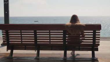 Uzun saçlı genç bir kadın deniz kenarında ahşap bir bankta oturuyor. Kayıklar gidiyor.