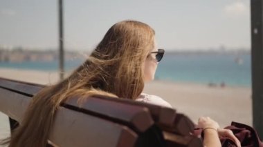 Genç bir kadın deniz kenarında bir bankta oturur ve uzaklara bakar, rüzgar uzun saçlarını dalgalandırır..