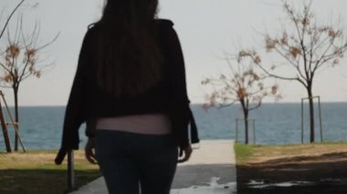 Genç bir kadın parkta denize giden bir yol boyunca yürür. Güneşli