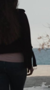 Genç bir kadın parkta denize giden bir yol boyunca yürür. Güneşli Dikey video.