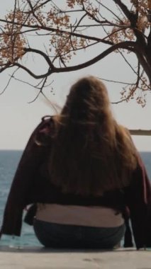 Genç bir kadın deniz kenarında betonun üzerinde oturur ve uzaklığa bakar. Rüzgar onun uzun saçlarını dalgalandırır. Dikey video.