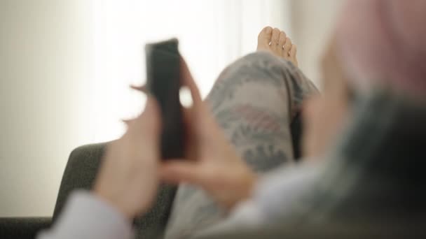 一个洗澡后的年轻女人正仰着腿躺在沙发上 头上拿毛巾手里拿着电话把重点放在腿上 — 图库视频影像