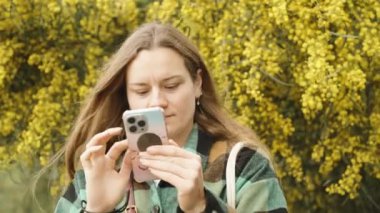 Genç bir kadın telefonuna sarı bir mimozanın arka planında bakıyor. Rüzgâr saçlarımı savuruyor.