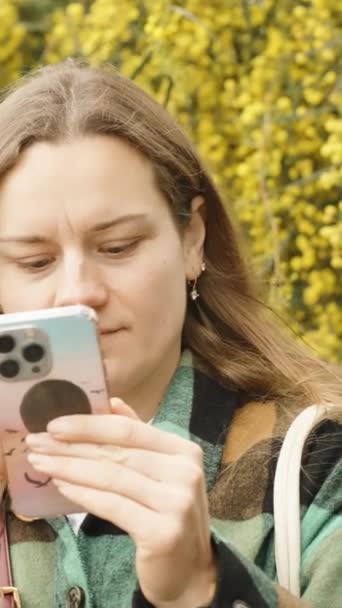 若い女性は 咲く黄色い模倣物の背景に対して彼女の携帯電話を見ます バーティカル — ストック動画