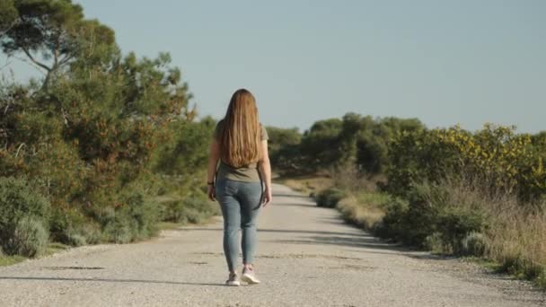 一个穿着牛仔裤和绿色T恤的年轻女子走在森林里一条废弃的铺面公路上 夕阳西下 后视镜 — 图库视频影像
