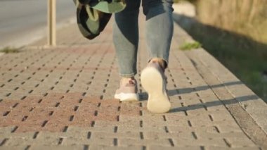 Kotlu ve elinde ekose gömleği olan genç bir kadın fayans yolu boyunca yürüyor, dikiz manzaralı. Cebinde bir telefon var. Batan güneş.