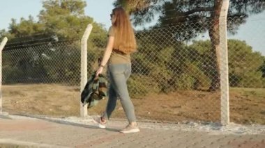 Güneşli bir akşamda dikenli tellerle çit boyunca yürüyen genç bir kadın, arkasında bir orman var..