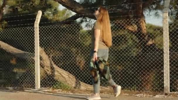 若い女性はメッシュと有刺鉄線で作られたフェンスで囲まれた松の森の近くを歩きます — ストック動画
