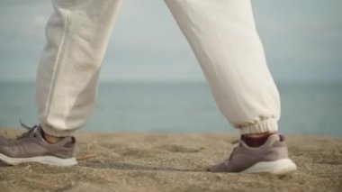 Mor spor ayakkabılı ayaklar ve beyaz pantolonlar deniz boyunca kumlu bir sahil boyunca yürüyorlar, bulutlu, yavaş.
