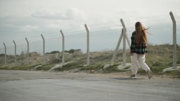若い女性はメッシュと有刺鉄線で作られたフェンスに沿って歩きます 砂漠の曇った天気 ゆっくりとした動き — ストック動画
