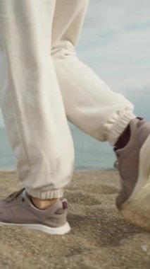 Dikey video. Genç bir kadın, bulutlu ve rüzgarlı havada, yavaş çekimde deniz kenarında yürüyor. Kamera geri gidiyor..