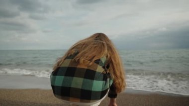 Genç bir kadın kumların üzerinde deniz kenarında oturuyor, soğuk rüzgarlı hava, kuma dokunuyor..