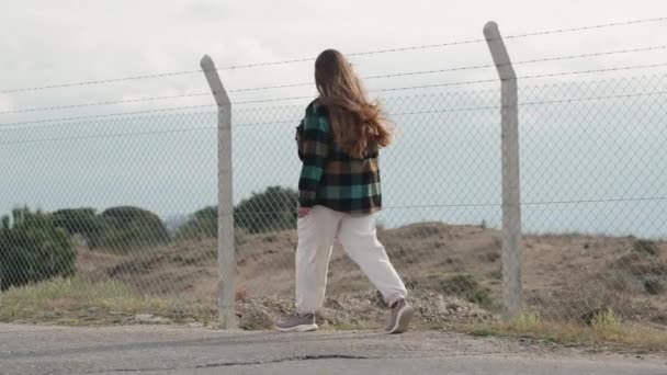 一个年轻的女人沿着公路走着 沿着一个铁丝网密布的铁丝网 多云多风的天气 吹拂着她的头发 篱笆后面有一片沙漠 — 图库视频影像