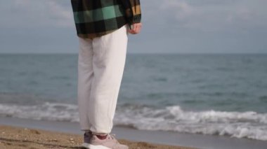 Genç bir kadın, bulutlu bir günde sıcacık giyinir deniz kenarına bakar, rüzgar saçlarını savurur..