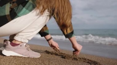 Genç bir kadın deniz dalgalarının yanında çömeliyor ve kuma dokunuyor..