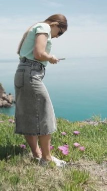 Kot etekli bir kız arka planda deniz olan yüksek bir tepede duruyor ve telefonuyla Carpobrotus 'un çiçeklerinin fotoğrafını çekiyor. Dikey video.