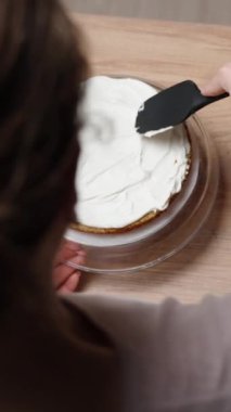 Kamera, pastayı hazırlarken kadın pasta şefinin arkasından ve üstünden çekim yapıyor. Sünger kekine krema sürüyor. Dikey video.