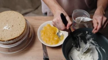 Bir kadın pasta hazırlıyor, kremadan bir kaşık temizliyor ve masada bisküvi ve turuncu dolgu var..