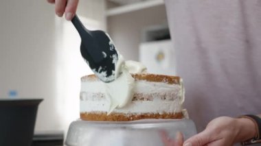 Kadın, spatula kullanarak pastanın kenarlarına beyaz krema sürüyor..