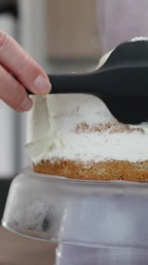 Bir kadın siyah spatula kullanarak bisküvi katmanlarına beyaz krema sürüyor. Pasta hazırlığı. Dikey video.