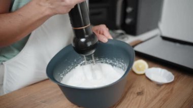 Ağır çekimde, bir kadın mavi bir kasede el çırpıcı kullanarak beyaz krema çırpmaya başlar..