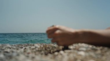 Denize yakın bir çakıl taşlı plajda bir kadın el, elindeki taşları topluyor ve denizin arka planına serpiyor..