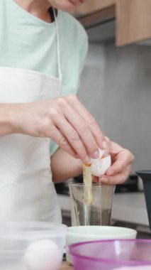 Parlak bir mutfakta bir kadın yemek pişiriyor. Yumurtayı kırıyor ve yumurta beyazını sarıdan bir bardağa ayırıyor. Dikey video.