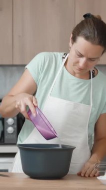 Genç bir kadın parlak bir mutfakta yemek pişiriyor. Tahta masadaki bir kaseye şeker döker. Dikey video.