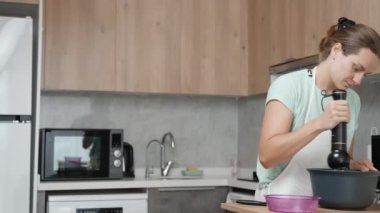 Mutfağın panoramik görüntüsüne göre beyaz önlüklü genç bir kadın mikserle malzemeleri çırpıyor..