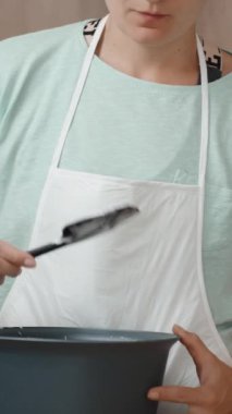 Beyaz önlüklü genç bir kadın mutfakta yemek pişiriyor ve spatulayı beyaz kremayla yalıyor. Dikey video.