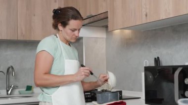 Beyaz önlüklü genç kadın parlak bir mutfakta yemek pişiriyor. Tabaktan tencereye bir şeyler döker..